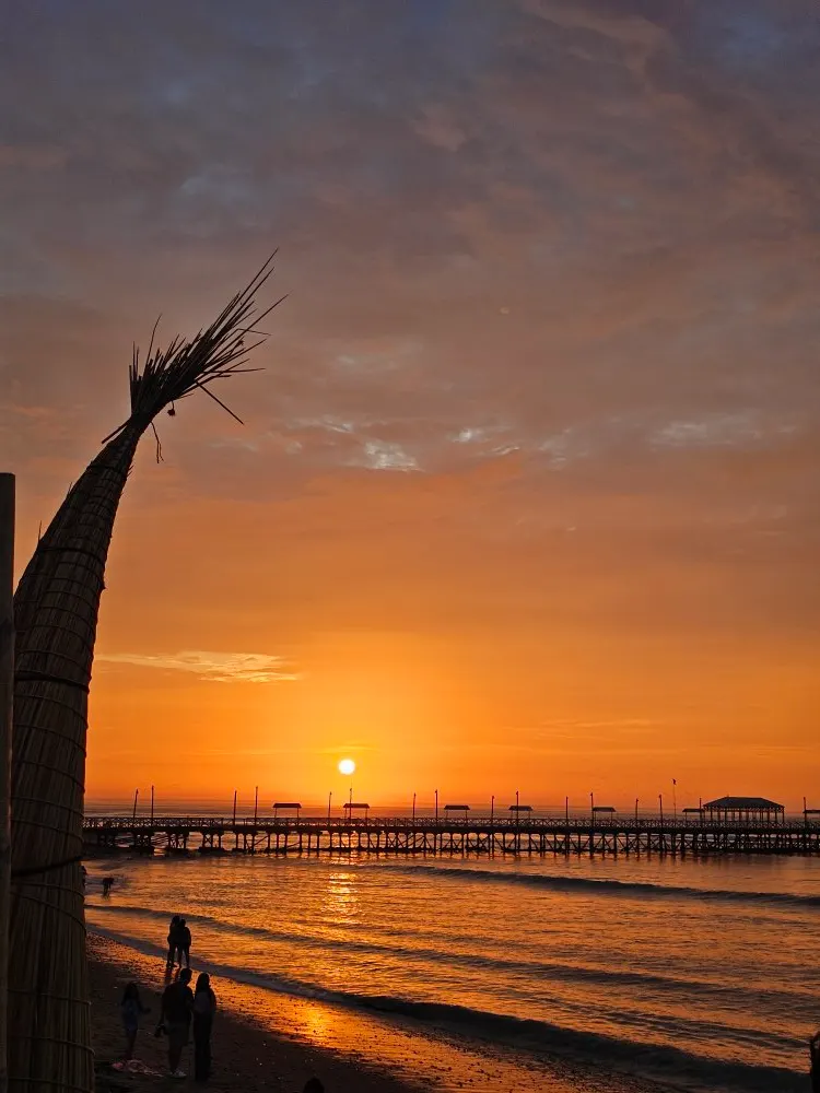 A long pier over the sea at sunset, with a traditional reed boat in the foreground